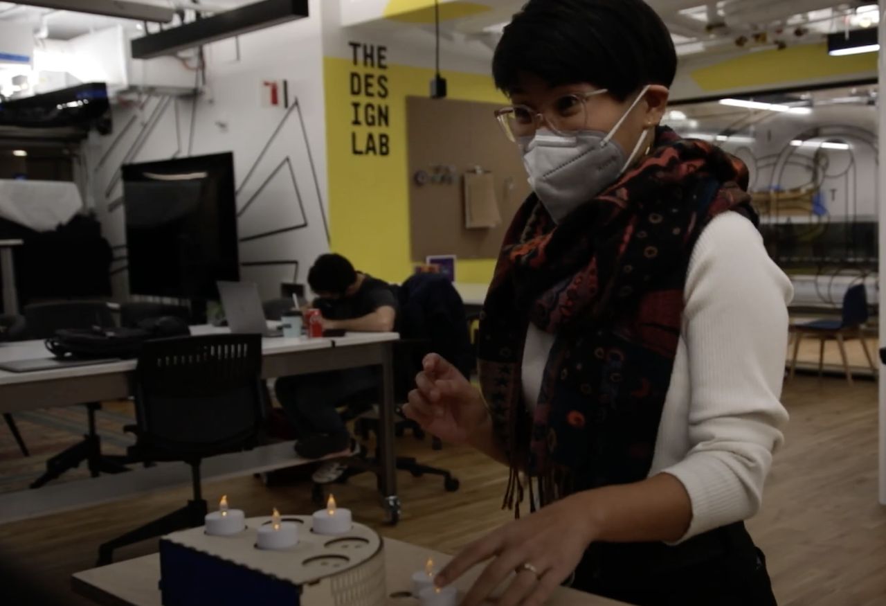 Image of an Asian person with short hare and a red patterned scarf. They are reaching down towards a table with fake candles on it, and looking to the left of the camera. They appear to be startled. In front of them is a candle holder with 3 candles. Behind them is a wall with "The Design Lab" written on it, and a person studying.