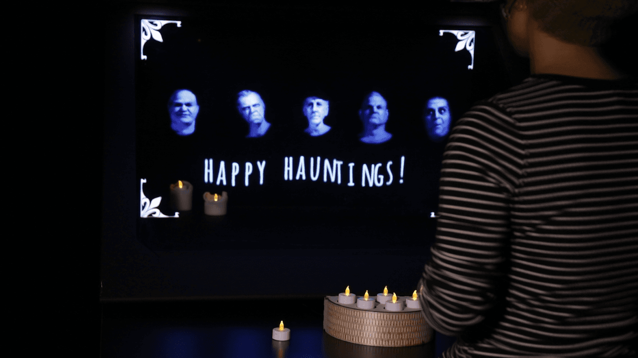 The scene is mostly black; on a table sits a wooden candle holder with 5 candles visibly on it. Behind it is a transparent screen with 5 ghostly heads hovering on it, over the words "Happy Hauntings!". Behind the transparent screen 2 candles are visible. Between the table and the camera is a person facing away from the camera.
