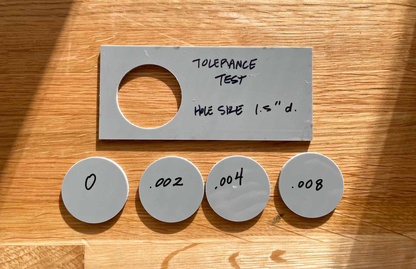 One large piece of acrylic has a circular hole. Four other circles of acrylic are below it on the table, each with a label on it detailing amount of tolerance given to the cut. Circles are labelled 0, .002, .004, .008.